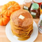 plate of pumpkin oat pancakes with butter and syrup on a white plate on a wooden counter next to spices and pumpkins.