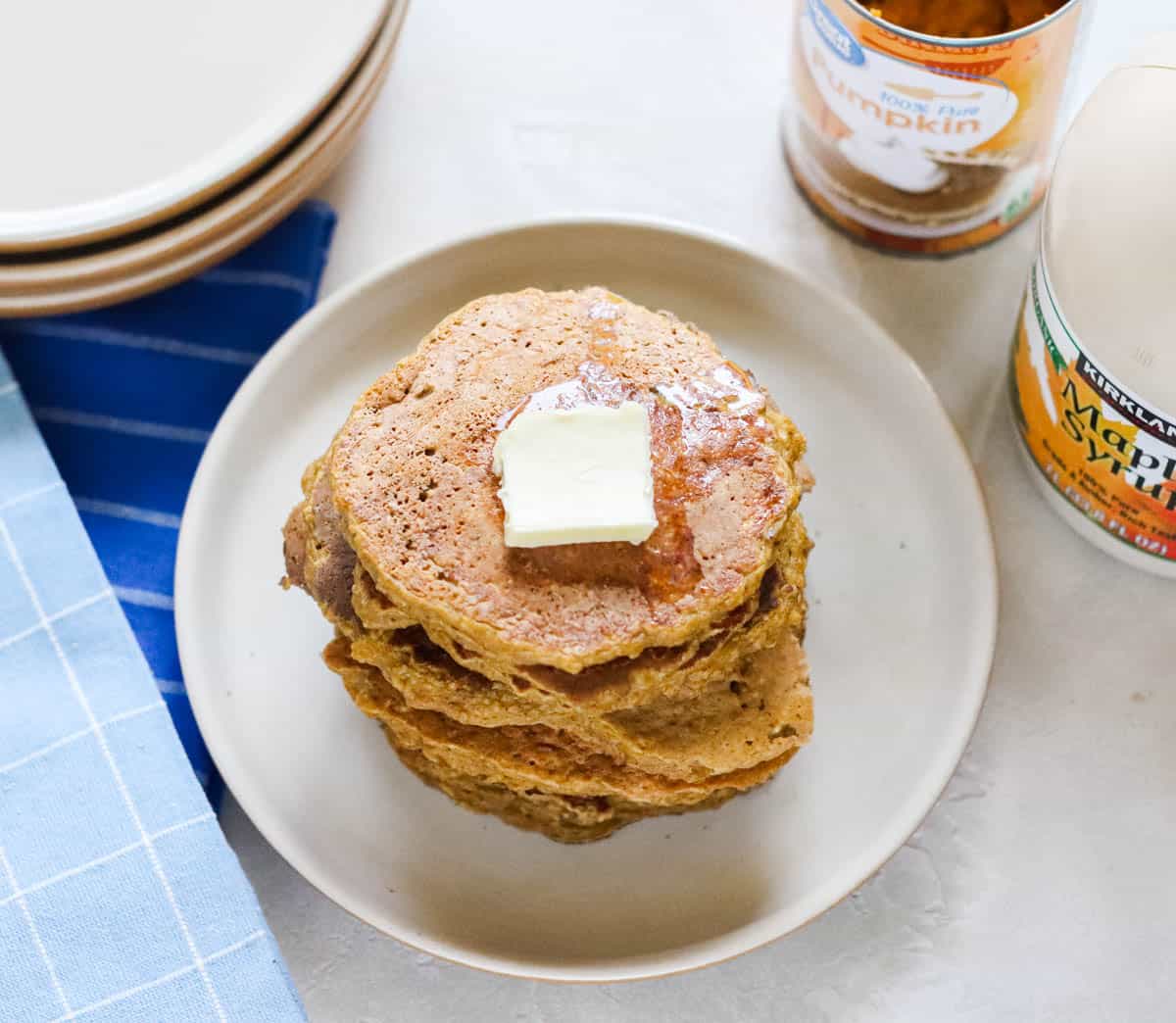 plate of pumpkin pancakes topped with butter next to blue napkins, a stack of plates, a bottle of maple syrup, and a can of pumpkin puree.
