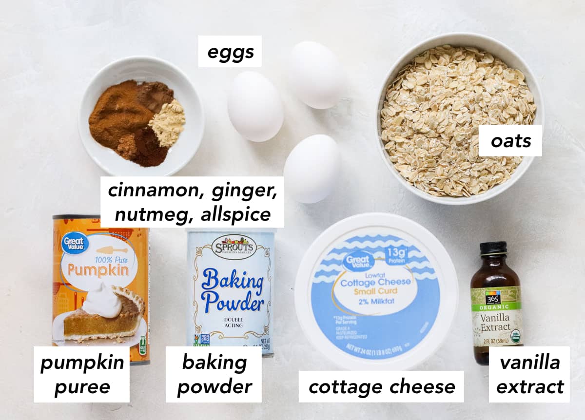 white counter with bowl of spices, 3 eggs, bowl of oats, can of pumpkin puree, can of baking powder, container of cottage cheese, and a bottle of vanilla extract with text overlay describing ingredients.