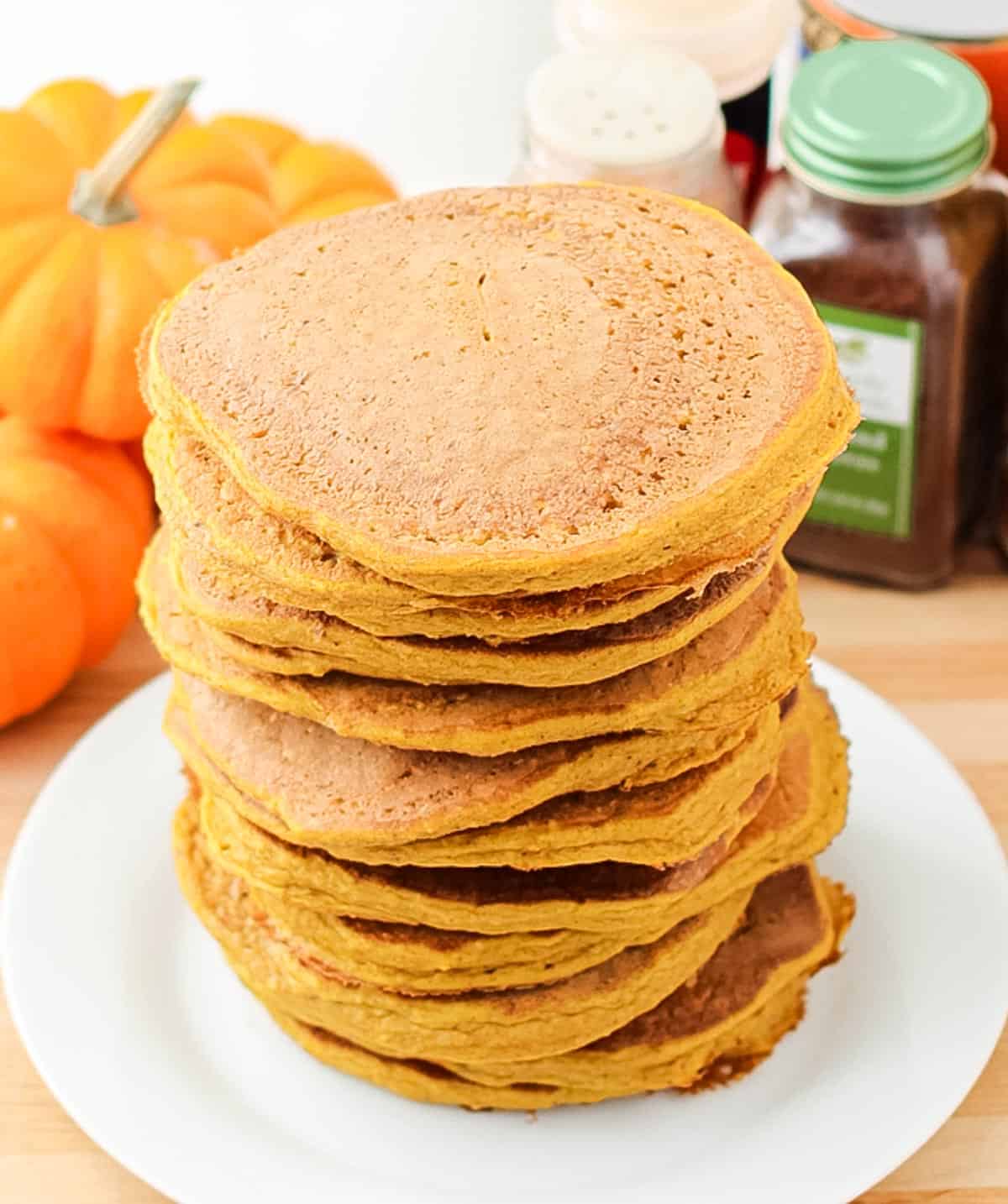 white plate with a stack of pumpkin pancakes surrounded by fresh pumpkins and spices.
