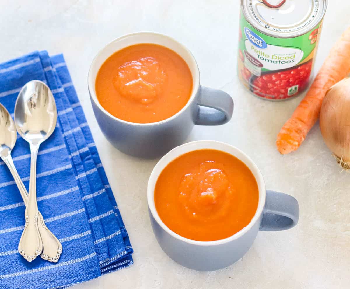 blue napkin with two spoons next to two mugs with tomato soup, a can of diced tomatoes, a carrot, and an onion.