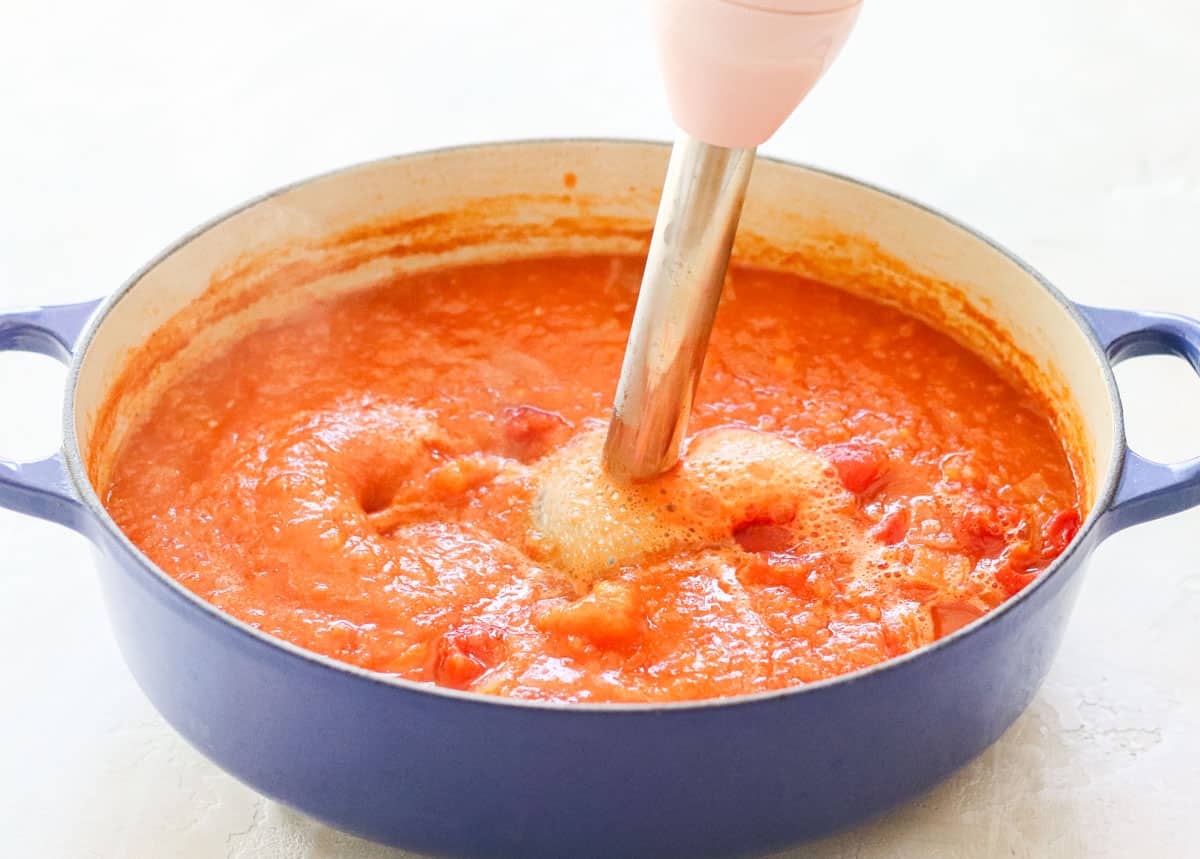 pink immersion blender blending together tomato soup in a blue pot.
