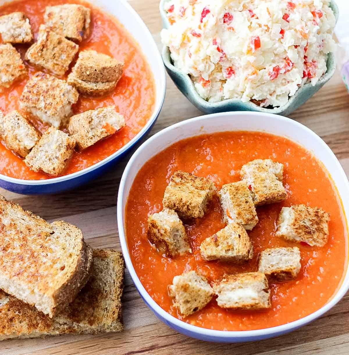 two bowls of tomato soup topped with grilled cheese cut into croutons next to a bowl of pimento cheese on a wooden cutting board.