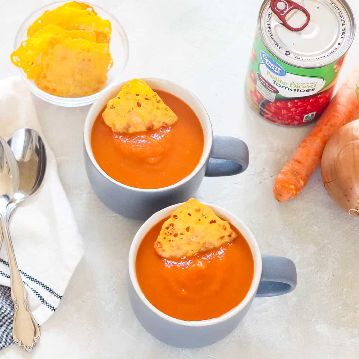 two mugs with tomato soup topped with cheese chips next to a napkin with two spoons, a carrot, a can of tomatoes, and an onion.