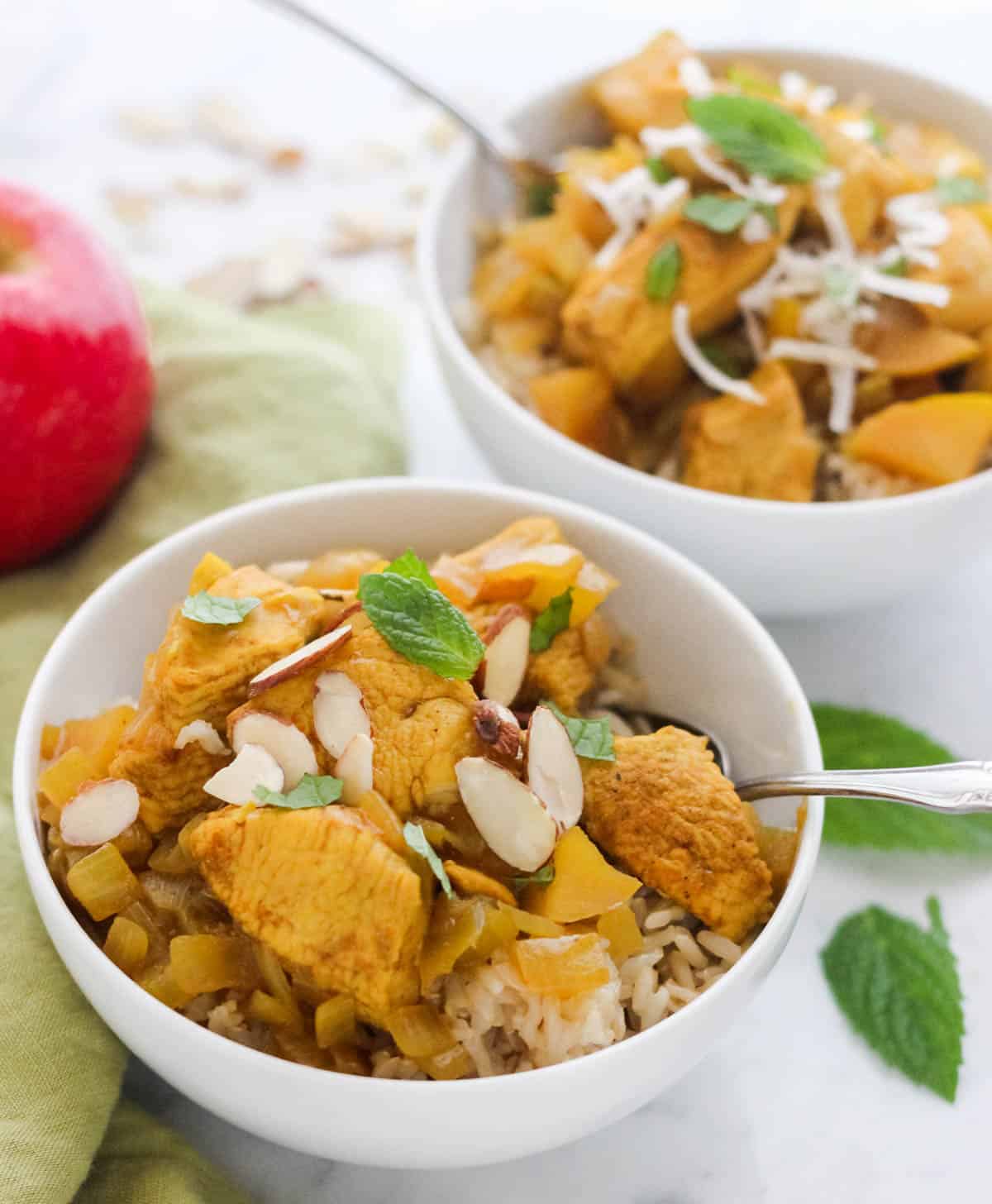 white bowl of healthy chicken curry topped with sliced almonds and fresh mint with silver spoons, another bowl in the background with an apple.