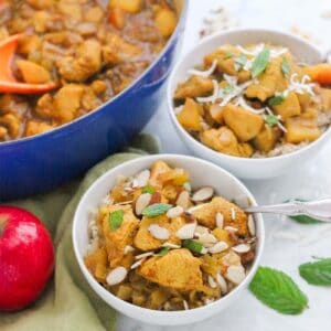 two white bowls of chicken apple curry topped with sliced almonds and fresh mint, apple, and blue pot with more apple curry sauce.