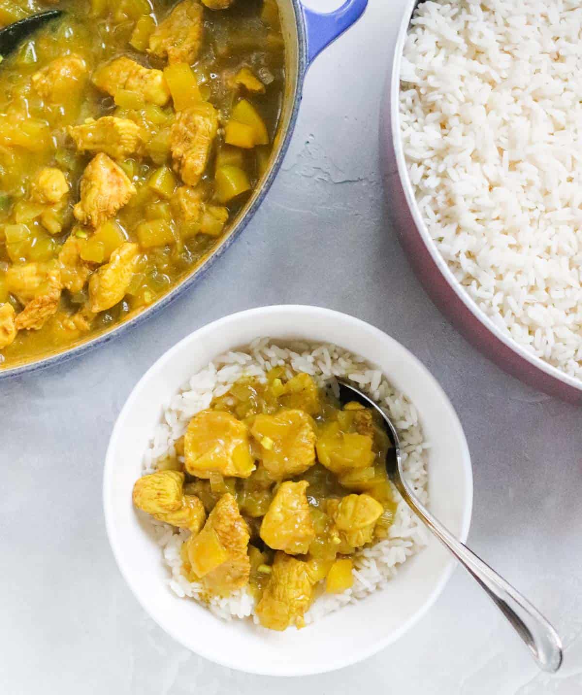 white bowl with rice and chicken apple curry with a silver spoon next to a blue pot of chicken curry sauce and a pot of white rice.