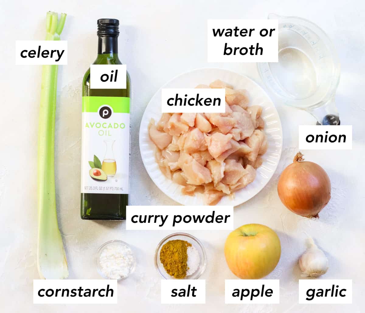 celery salt, bottle of avocado oil, bowl of cornstarch, plate of raw chicken, bowl of salt and curry powder, apple, head of garlic, yellow onion, and water in a measuring cup with text overlay describing ingredients.