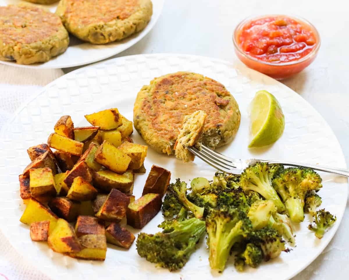 white plate with roasted broccoli and potatoes, lime slice, and avocado salmon burger with a fork holding a bite of the burger next to a plate of more burgers.