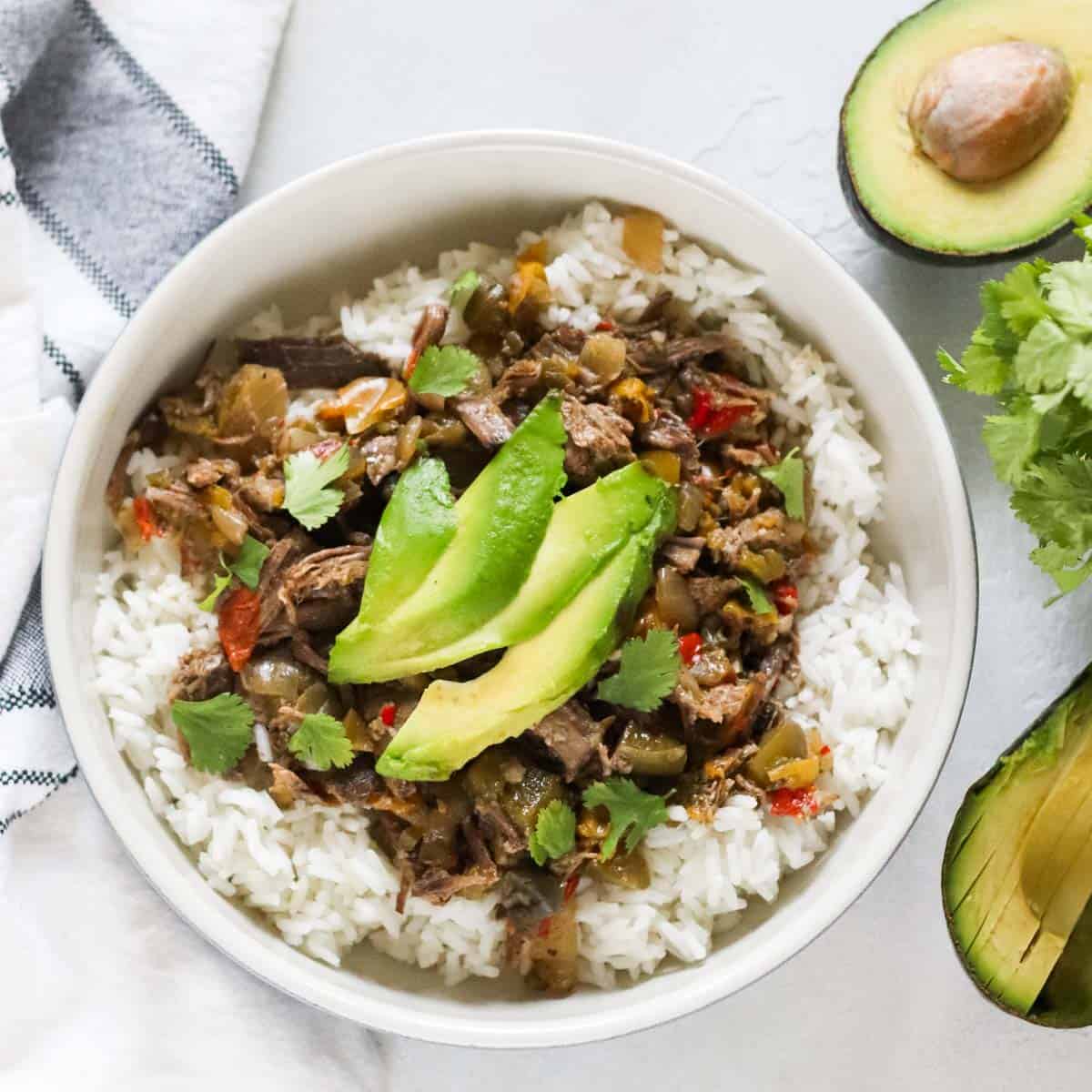 cut fresh avocado next to a bowl of rice with beef carnitas topped with sliced avocados.