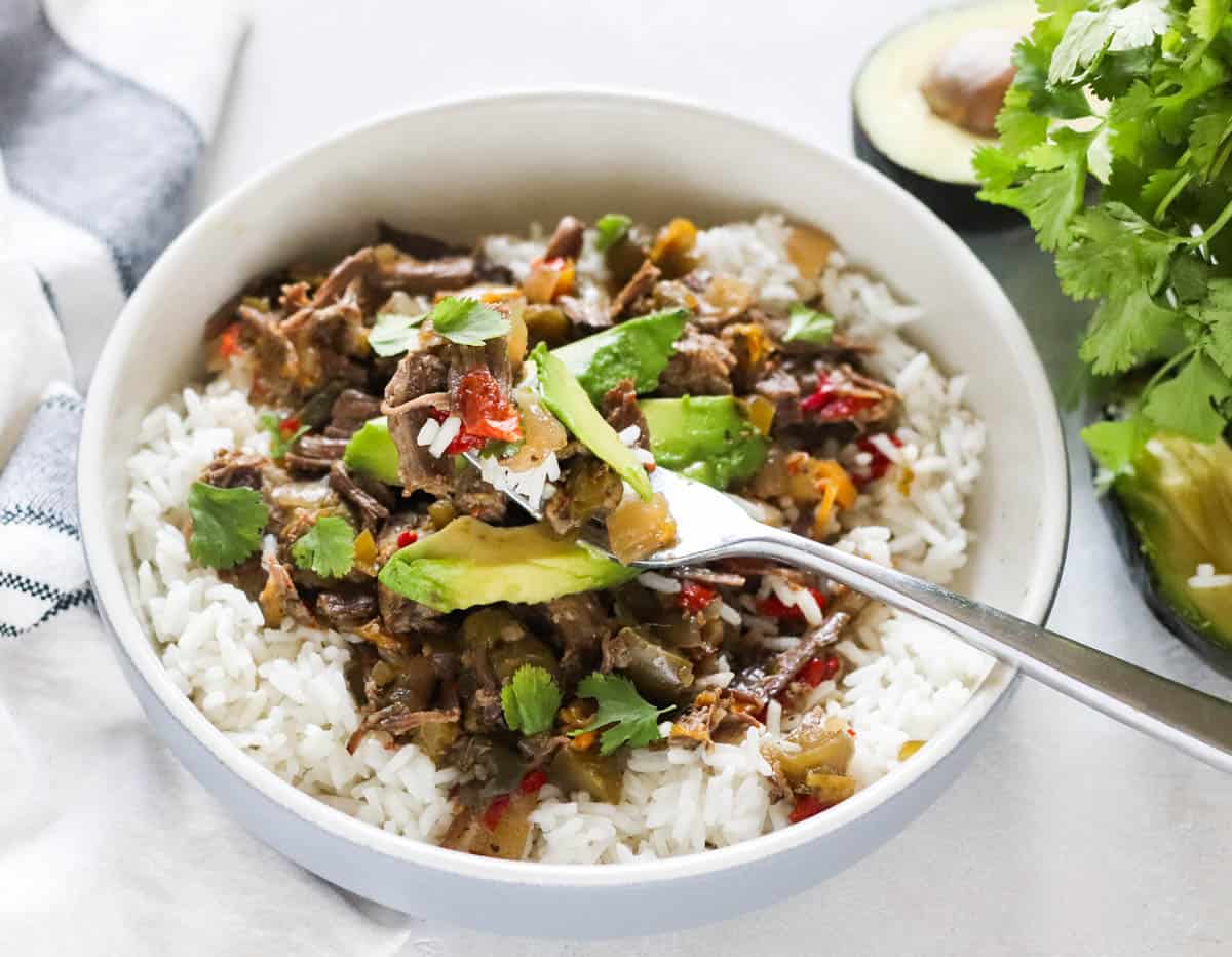 fork with beef carnitas and avocado over a bowl of rice with beef carnitas next to a white and black napkin.