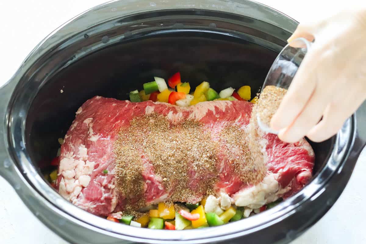 hand sprinkling a bowl of spices over skirt steak and veggies in a slow cooker.