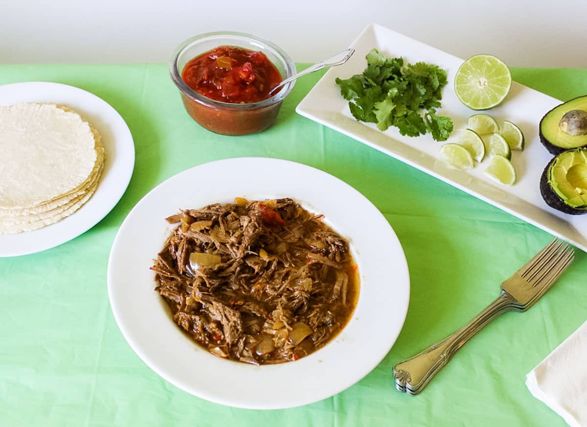 white plate with corn tortillas, a white bowl filled with beef baracoa, a bowl of salsa, and a white plate with avocado, lime slices, and cilantro.