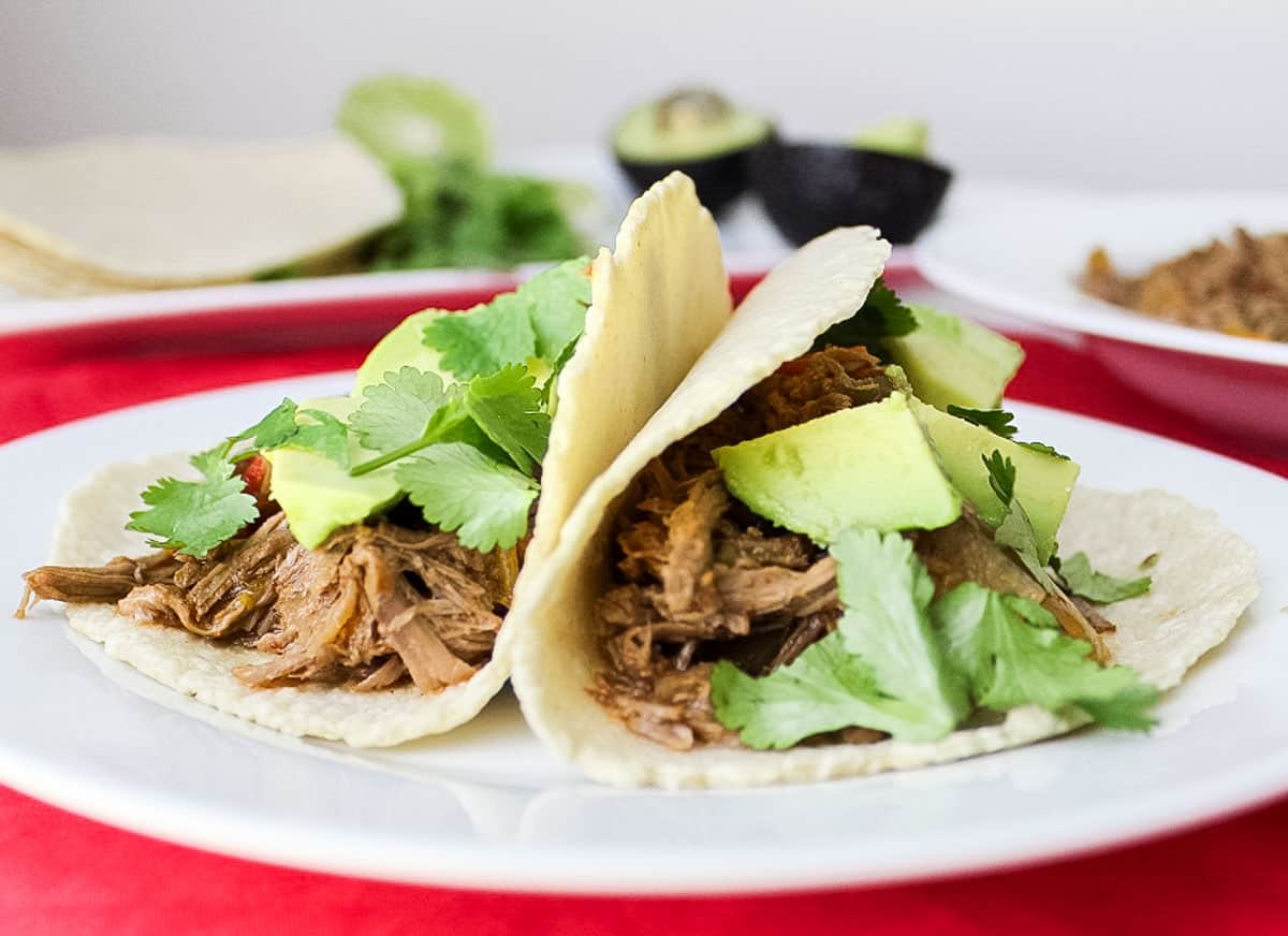 red tablecloth with two beef carnitas tacos on a white plate with avocado, cilantro, and lime slices.