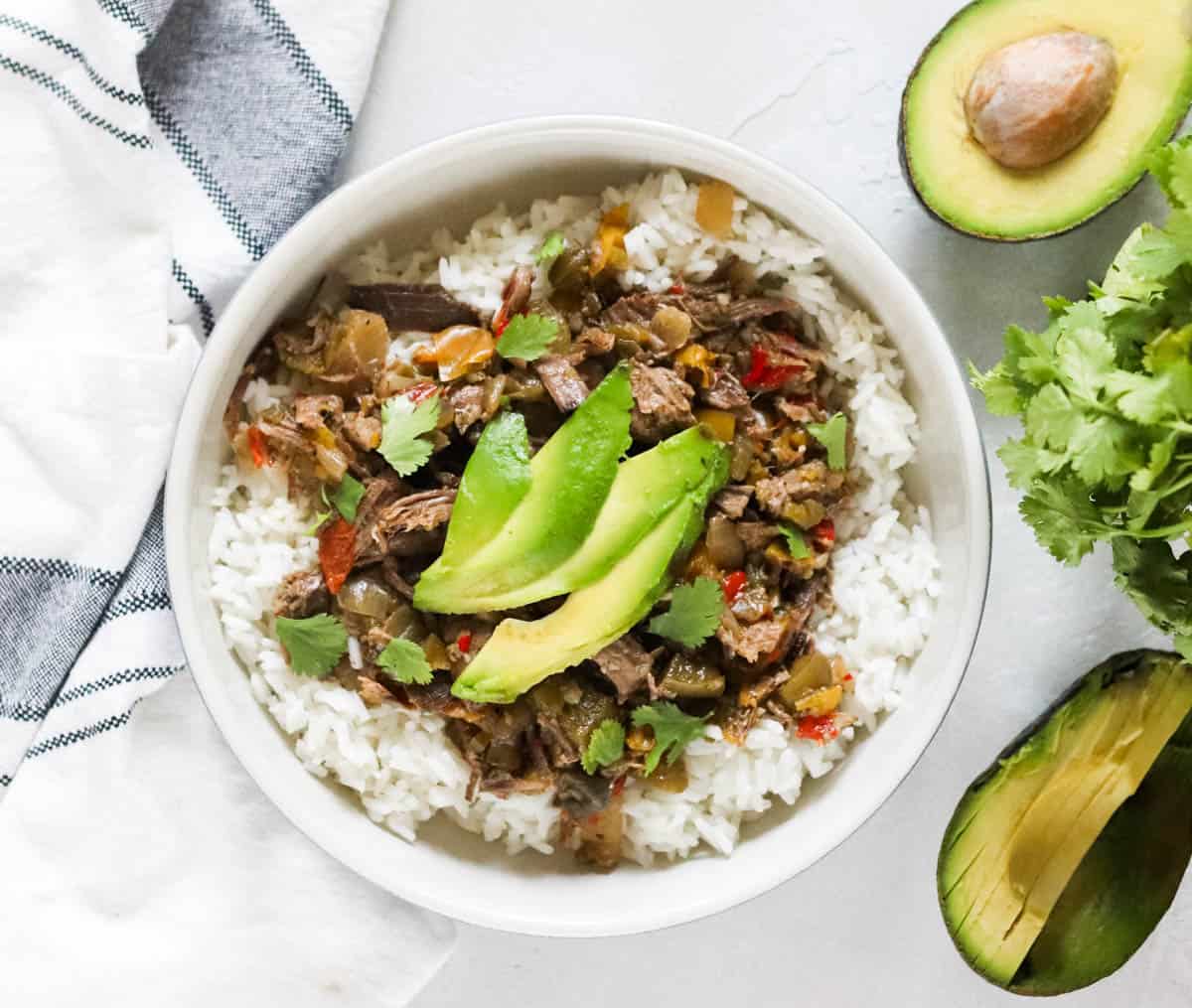 bowl of beef carnitas and rice topped with avocado slices next to a napkin and whole cut avocados.