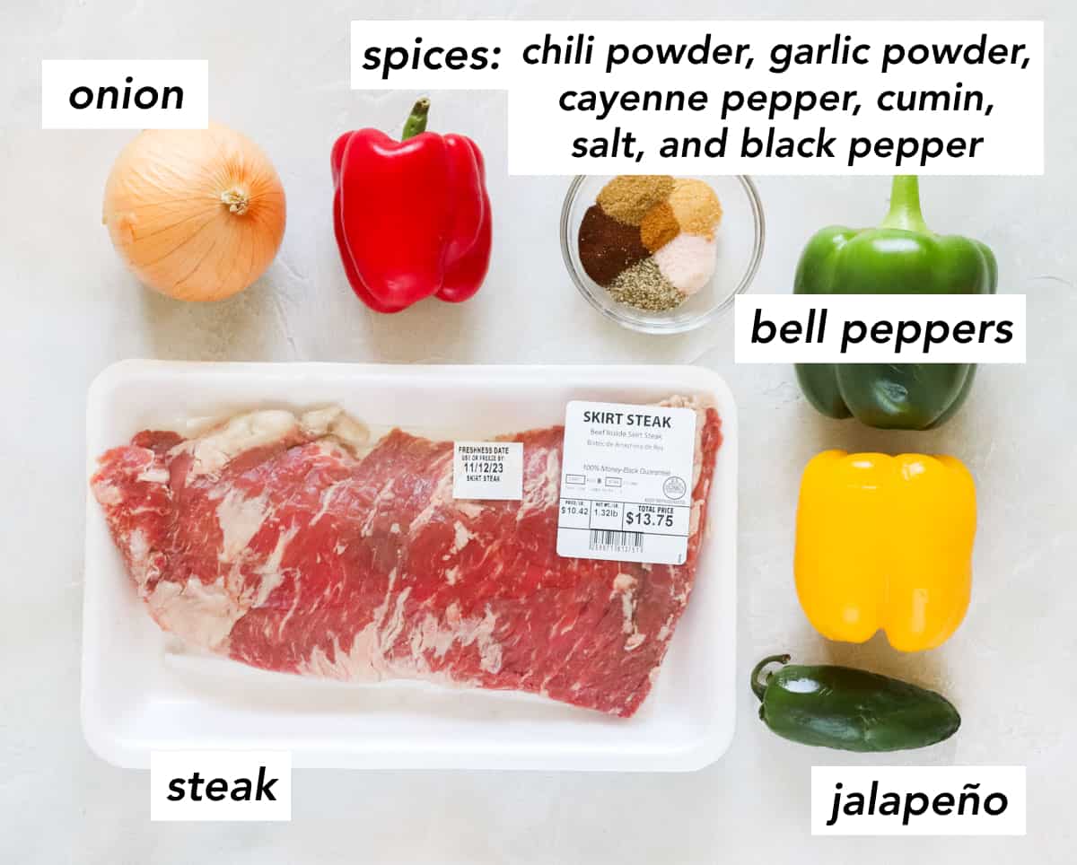 yellow onion, red bell pepper, skirt steak, jalapeno, yellow bell pepper, green bell pepper, and a bowl of spices on a white counter.