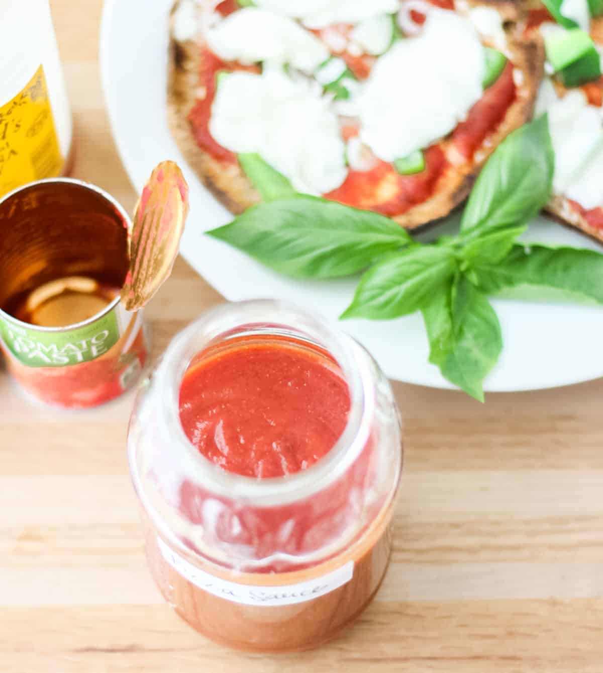 glass jar with homemade pizza sauce on a wooden table with an empty tomato paste can next to a plate with pita pizzas.