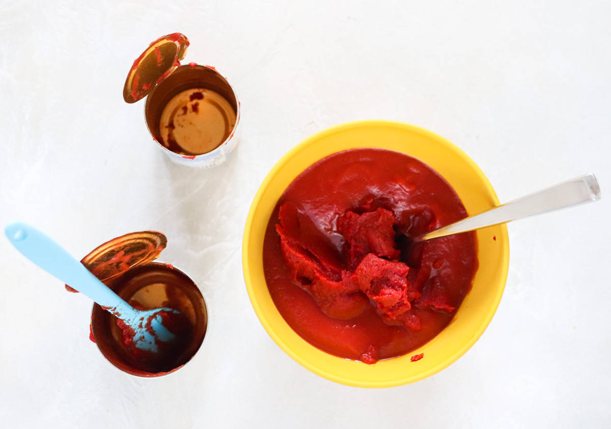 empty cans with a blue spatula next to a yellow bowl with tomato sauce and tomato paste.