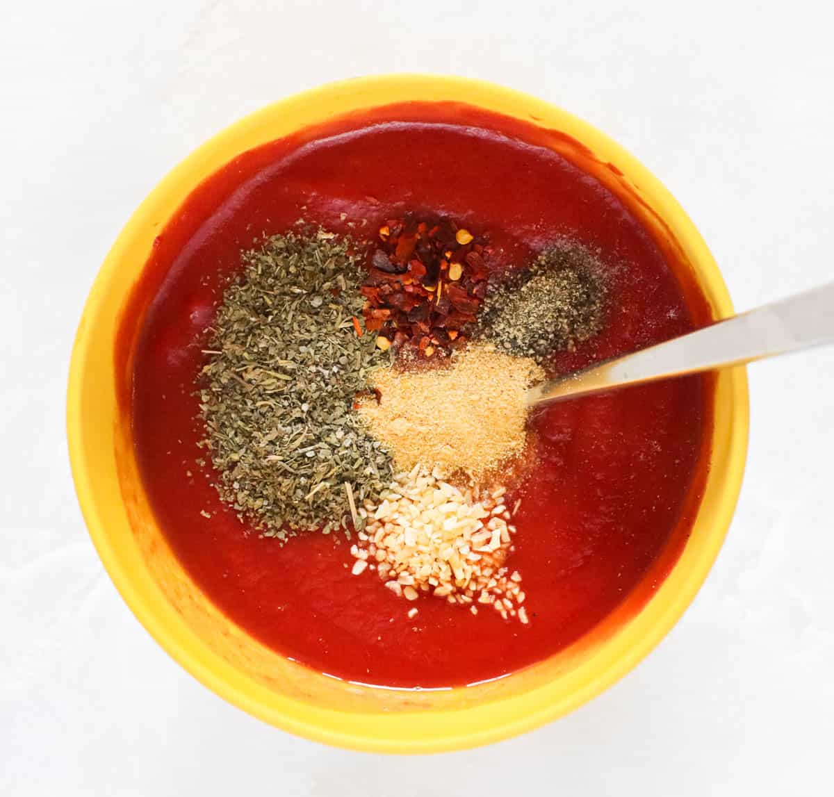yellow bowl with tomato sauce, tomato paste, onion flakes, garlic powder, black pepper, and red pepper flakes with a fork to stir.