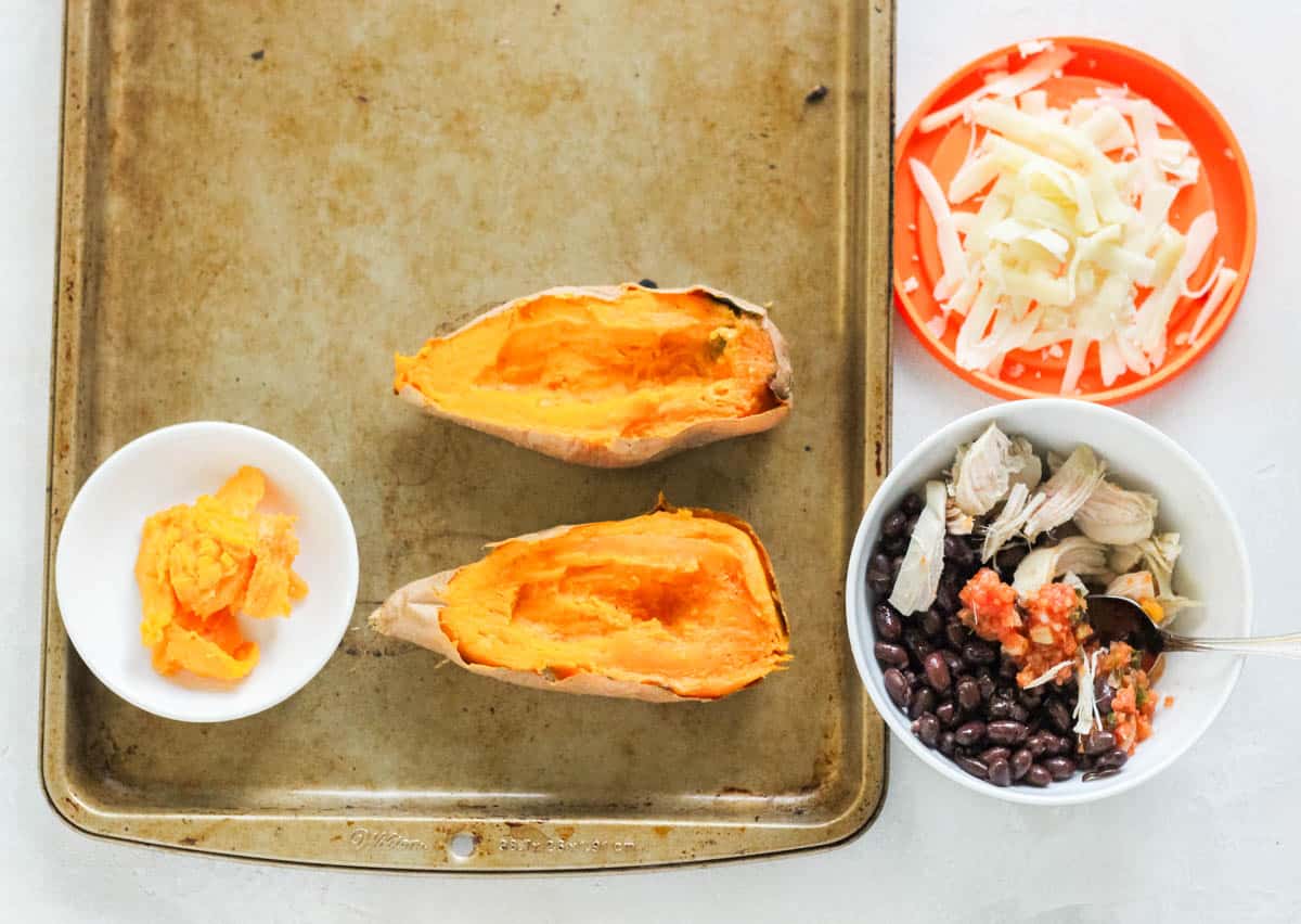 baking sheet with halved baked sweet potato and a bowl of sweet potato flesh, next to a bowl of chicken, salsa, and black beans being stirred by a silver spoon.