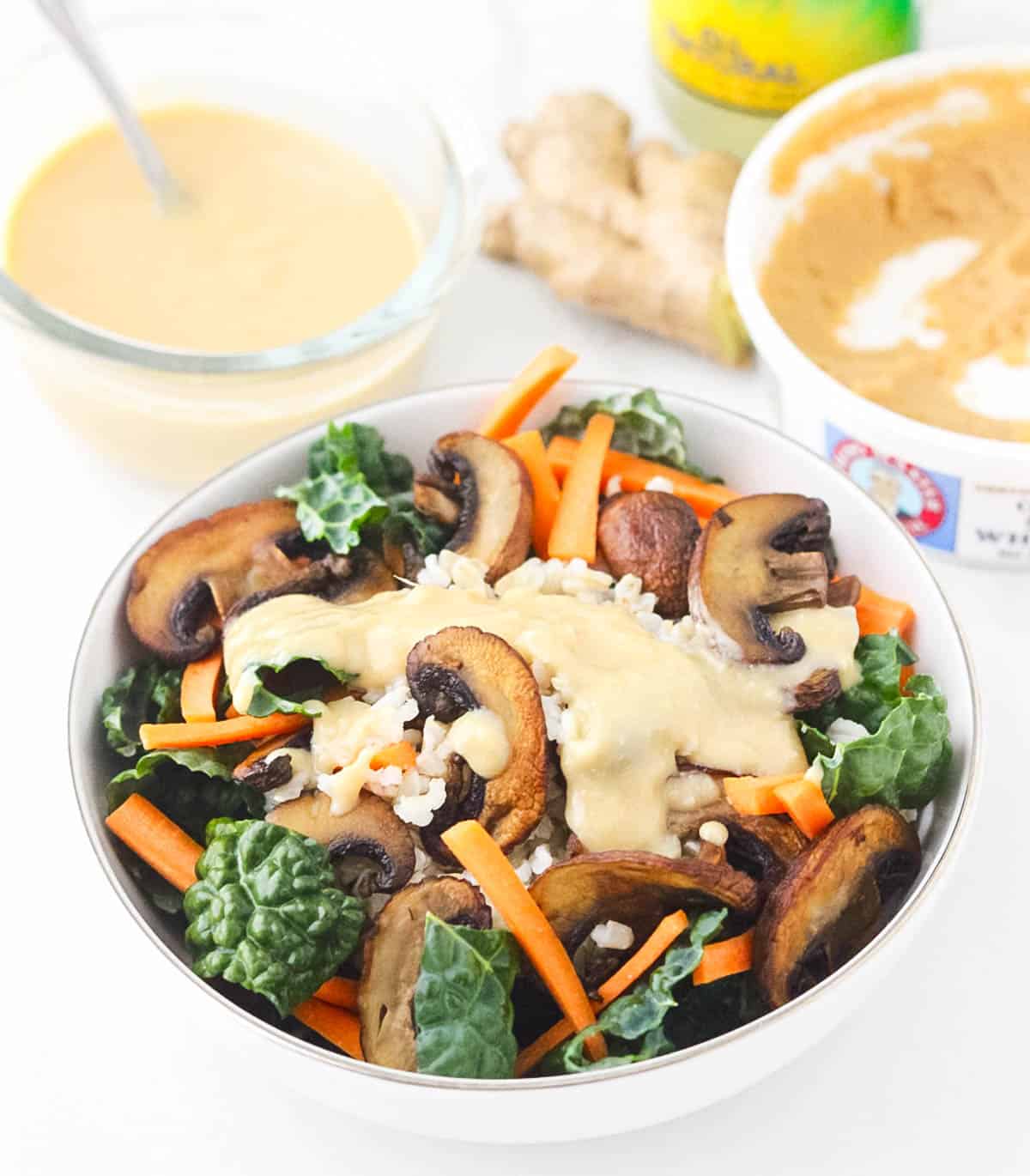 white bowl with kale, brown rice, carrots, and mushrooms topped with a miso dressing with miso, fresh ginger, and extra miso sauce in background.