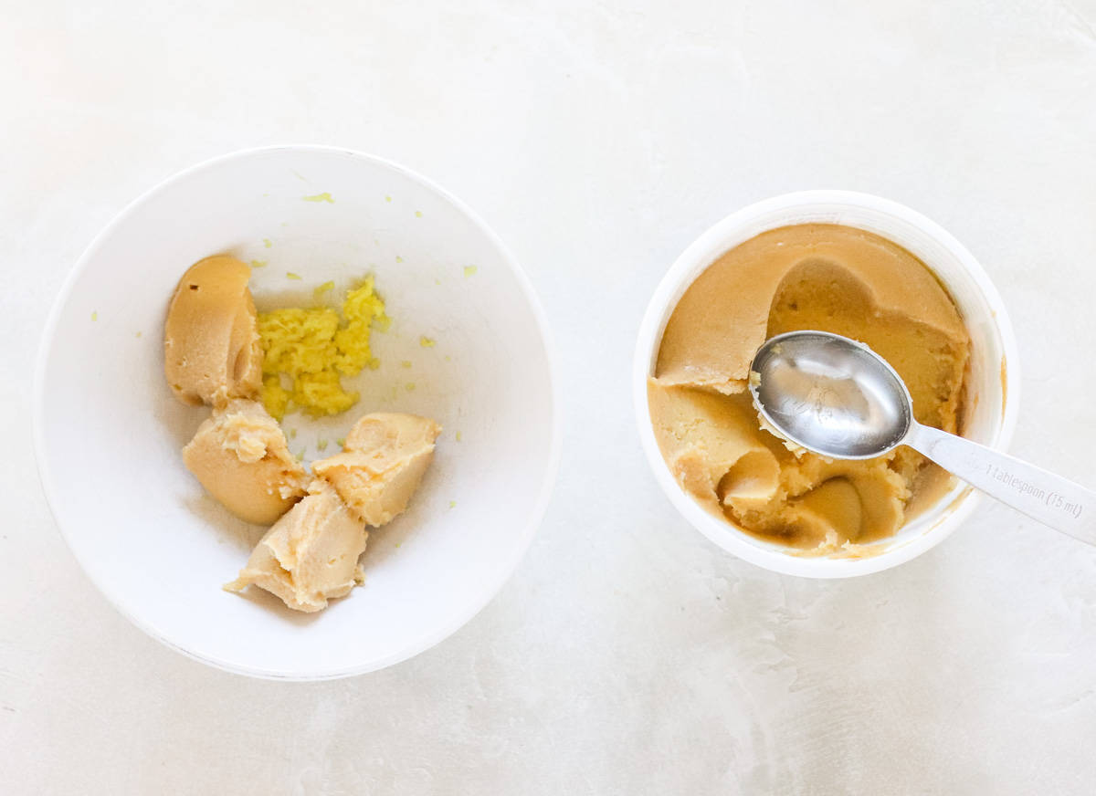 white bowl with white miso paste and grated ginger next to a container of miso paste with a silver measuring spoon.