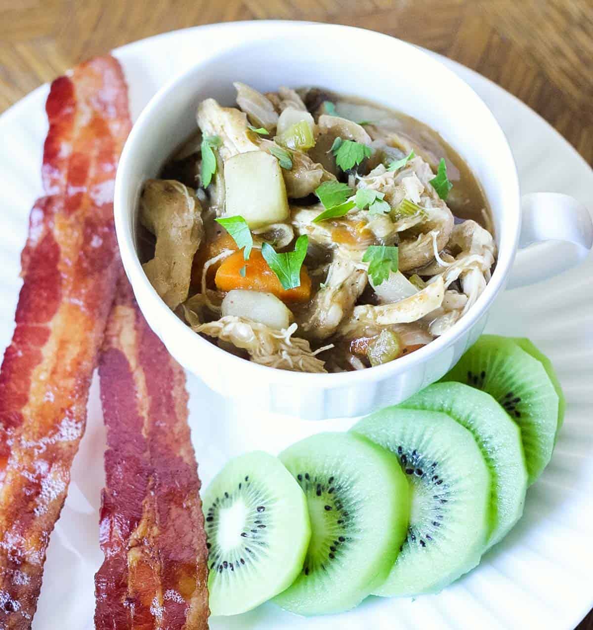 plate with bacon and sliced kiwi with a bowl of chicken hash.