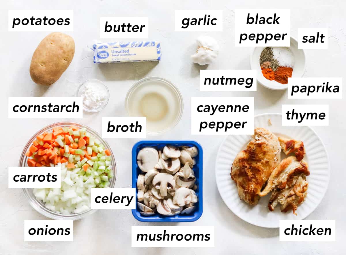 white counter with a russet potato, bowl of cornstarch, bowl of chicken broth, stick of butter, head of garlic, bowl of spicy, plate of cooked chicken breasts, container of sliced mushrooms, and a bowl of diced carrots, celery, and onions.
