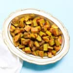 gold and white porcelain bowl filled with curry roasted potatoes next to a white napkin on a blue counter.