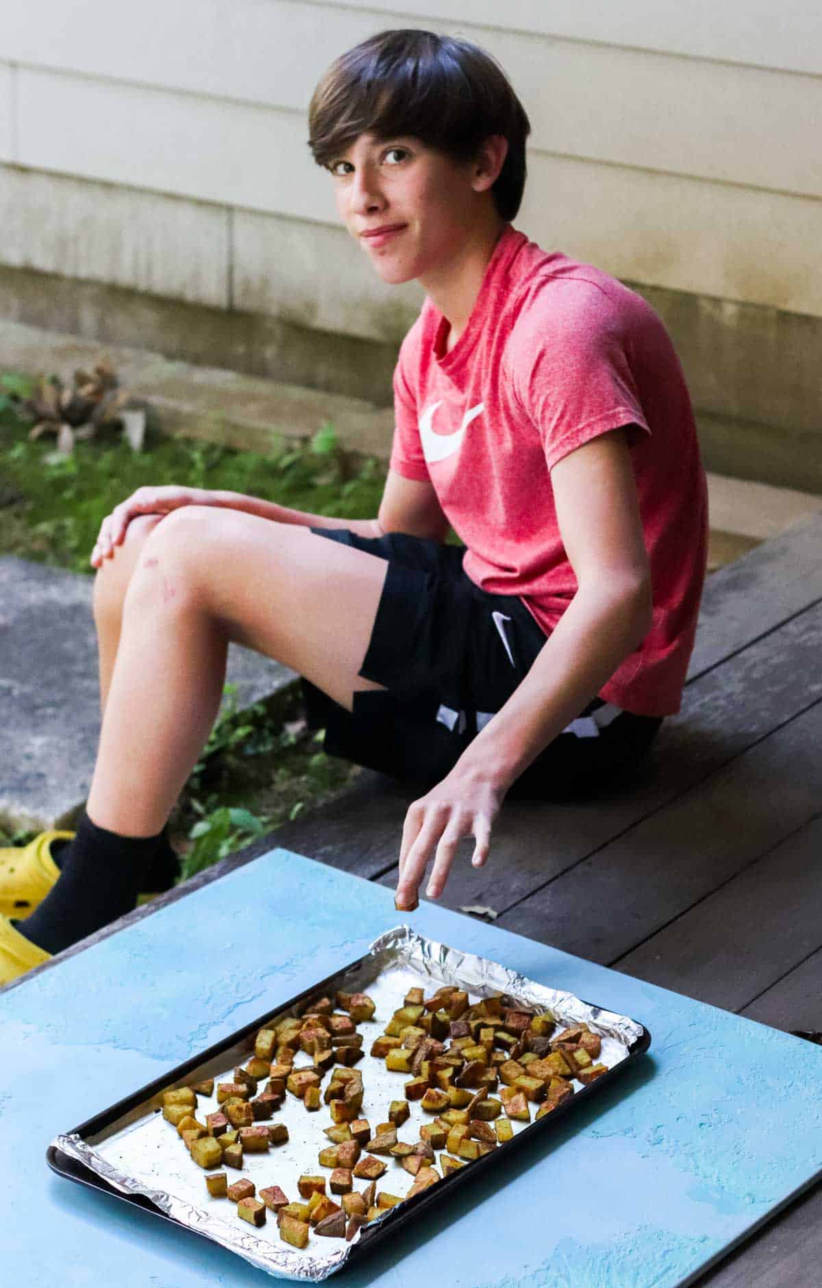 brunette teenage boy grabbing a roasted curry potato off the baking sheet sitting on a back porch.