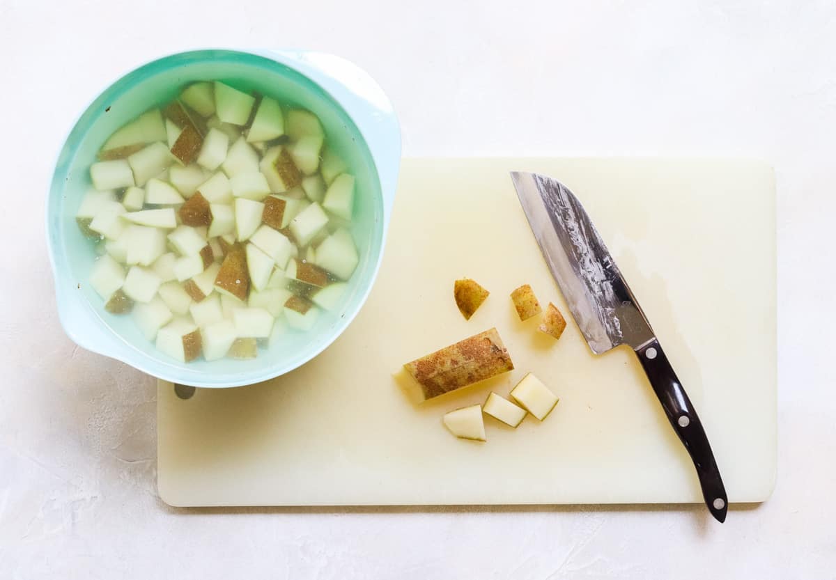 cutting board with knife chopping potatoes next to a blue mixing bowl with chopped potatoes soaking.
