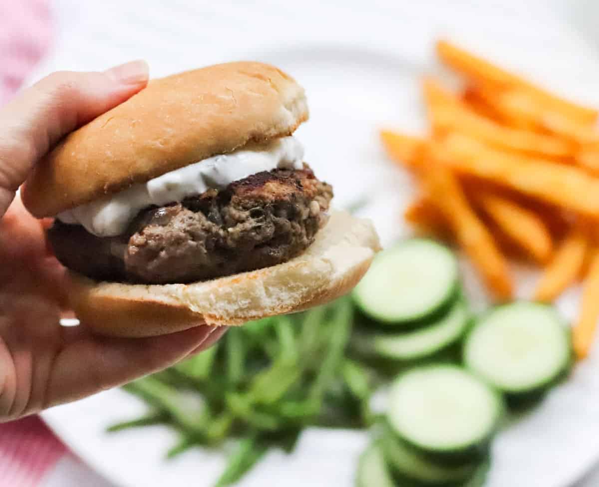 hand holding a lamb burger on a bun with tzatziki sauce over a plate of fries, green beans, and cucumbers.
