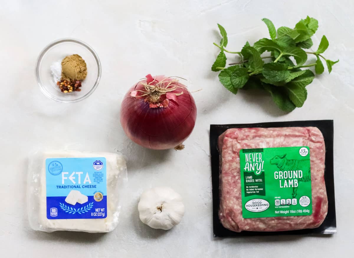 ingredients for lamb burgers on a white counter with ground lamb, feta cheese, garlic, red onion, bowl of spices, and fresh mint.