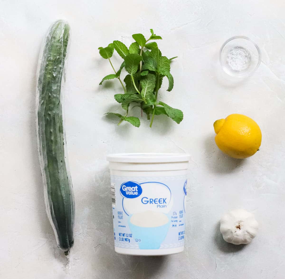 fresh cucumber, container of plain yogurt, fresh garlic, lemon, fresh mint, and a small bowl of salt on a white counter.