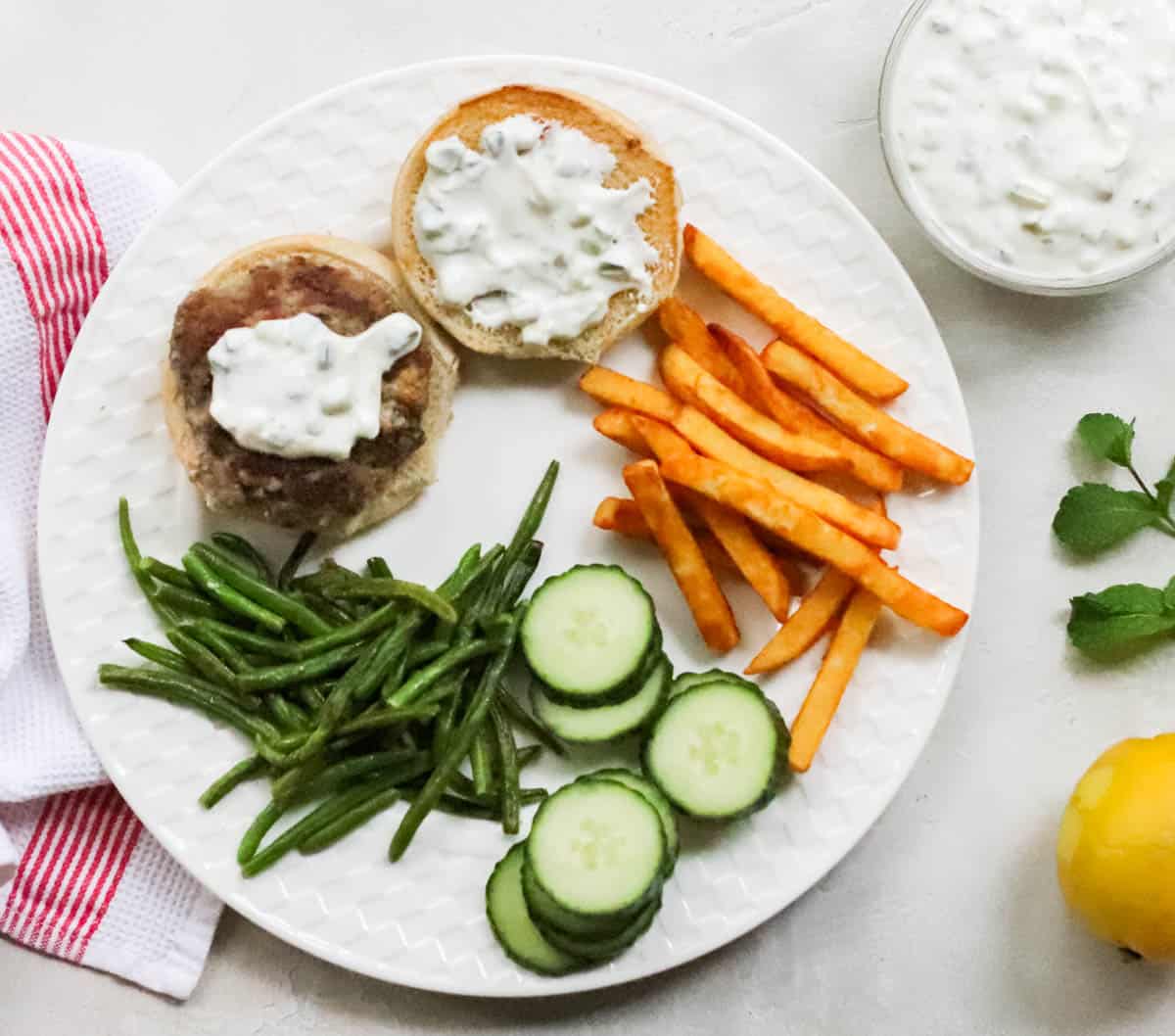 fresh mint and lemon next to a white plate with lamb burger, sliced cucumbers, tzatziki sauce, roasted green beans, and fries.