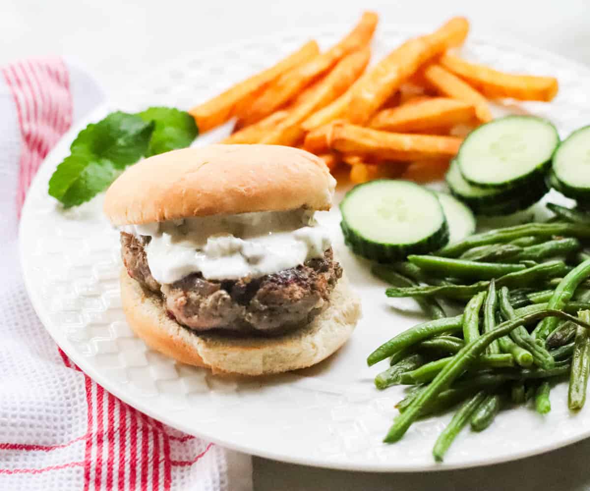 white plate with roasted green beans, sliced cucumber, fries, and a lamb burger.
