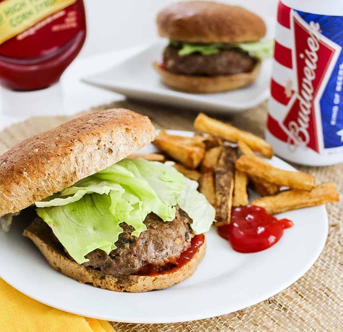 white plate with a burger topped with lettuce and a bun next to homemade fries, can of beer and extra ranch burger in background.