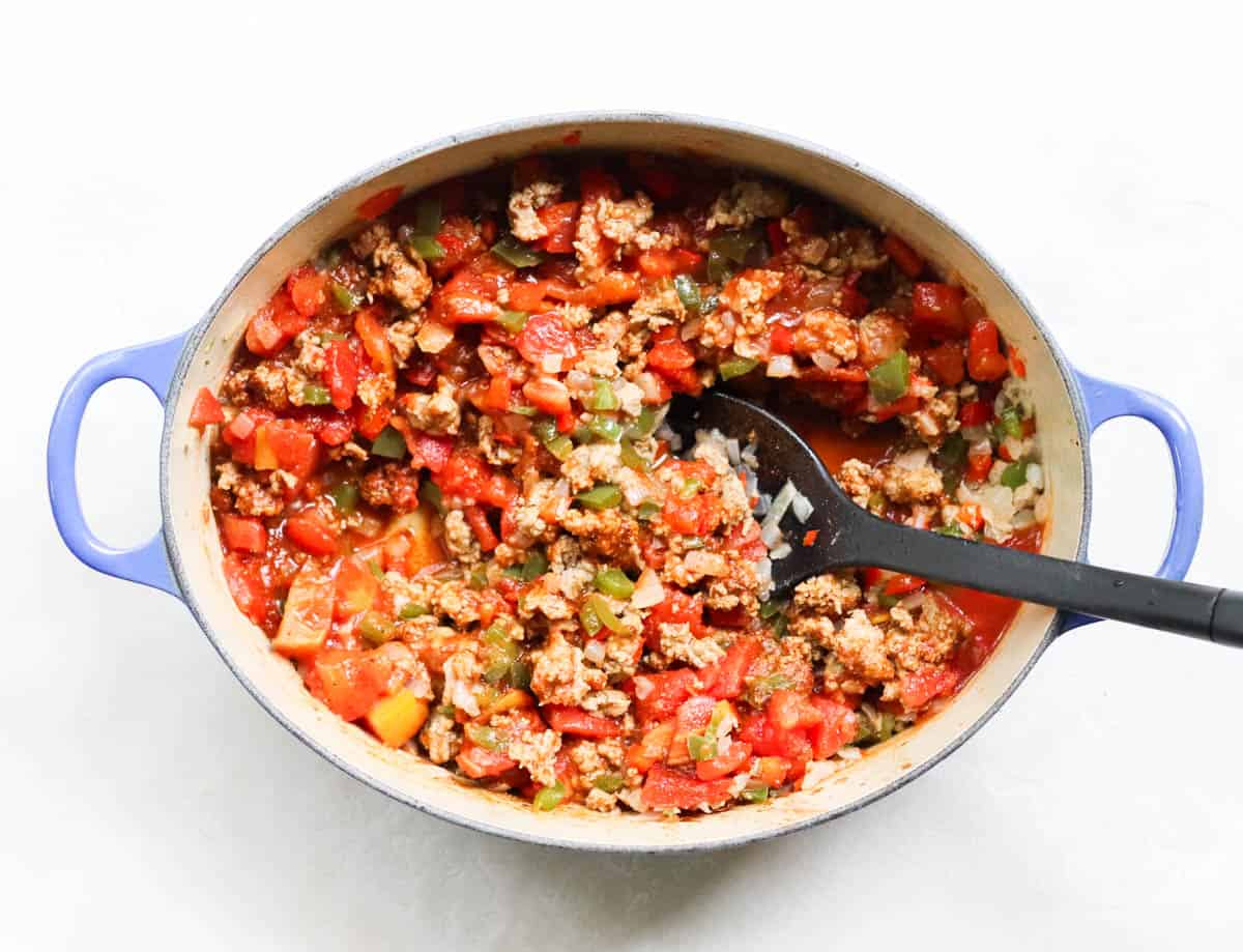blue pot with ground beef, veggies, and canned tomatoes stirred together with black spoon.