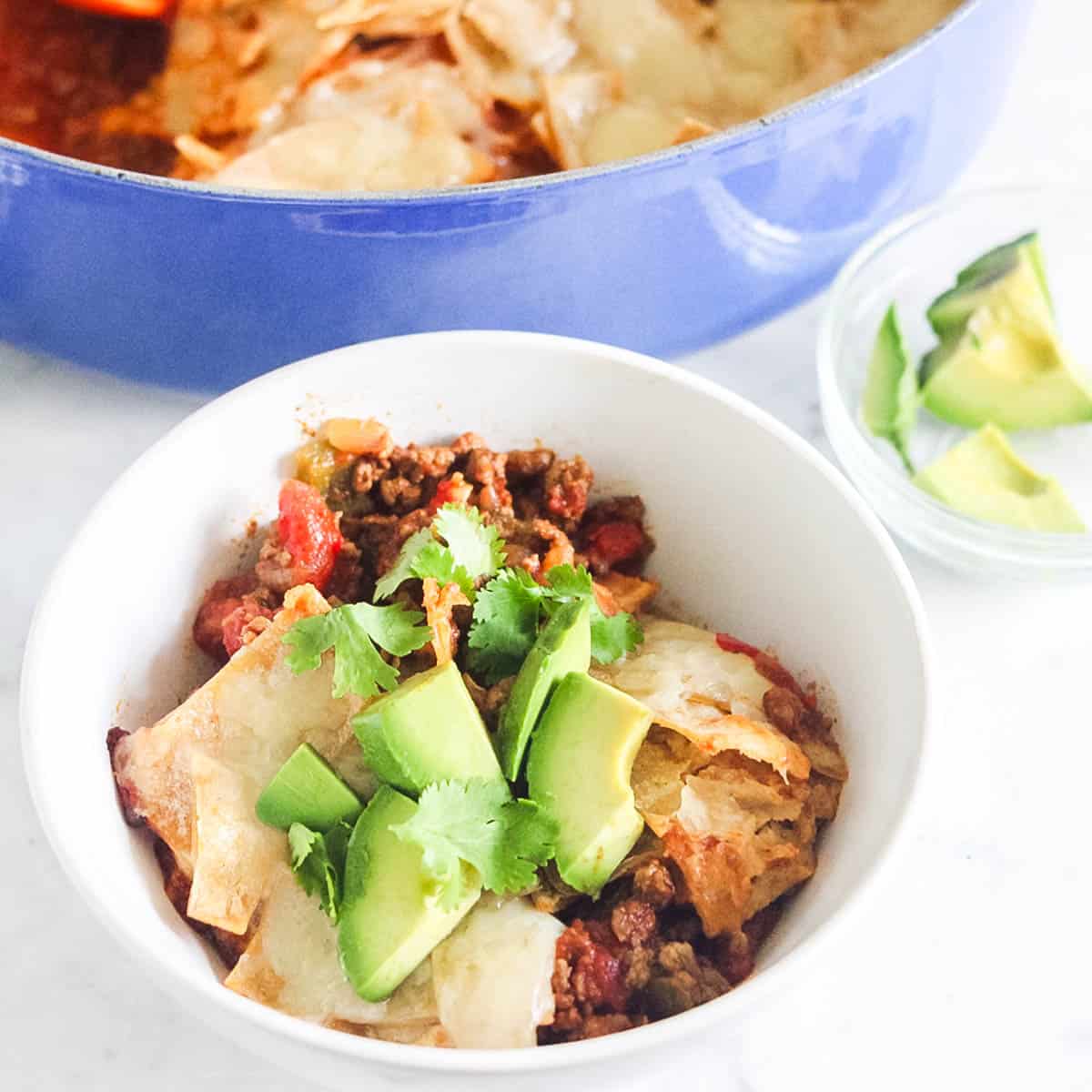 white bowl with taco casserole topped with cilantro and fresh avocado with a blue pot of taco casserole and bowl of avocado in the background.