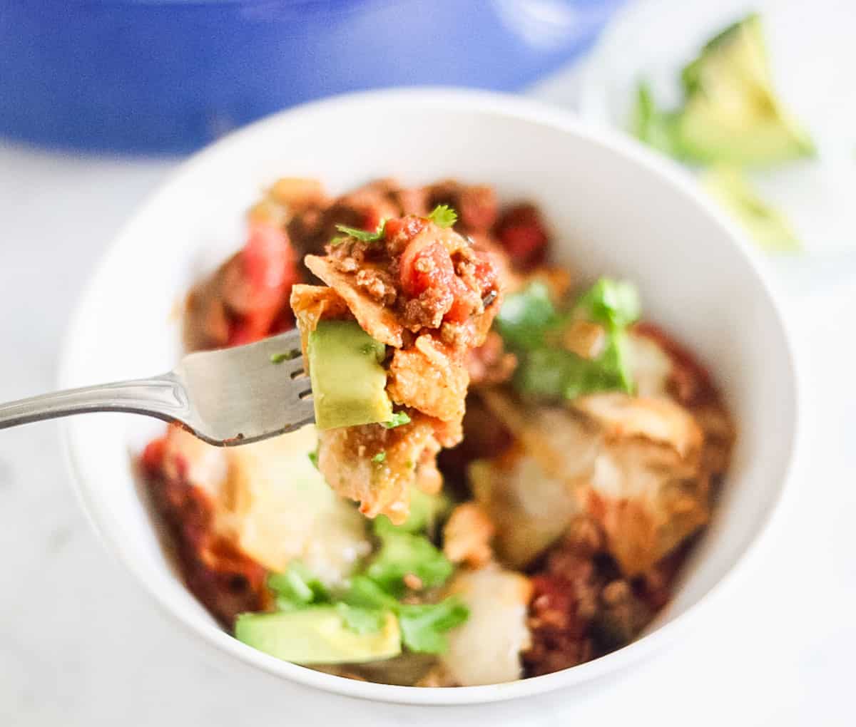fork holding a bite of taco casserole over a bowl of the casserole topped with avocado and cilantro.