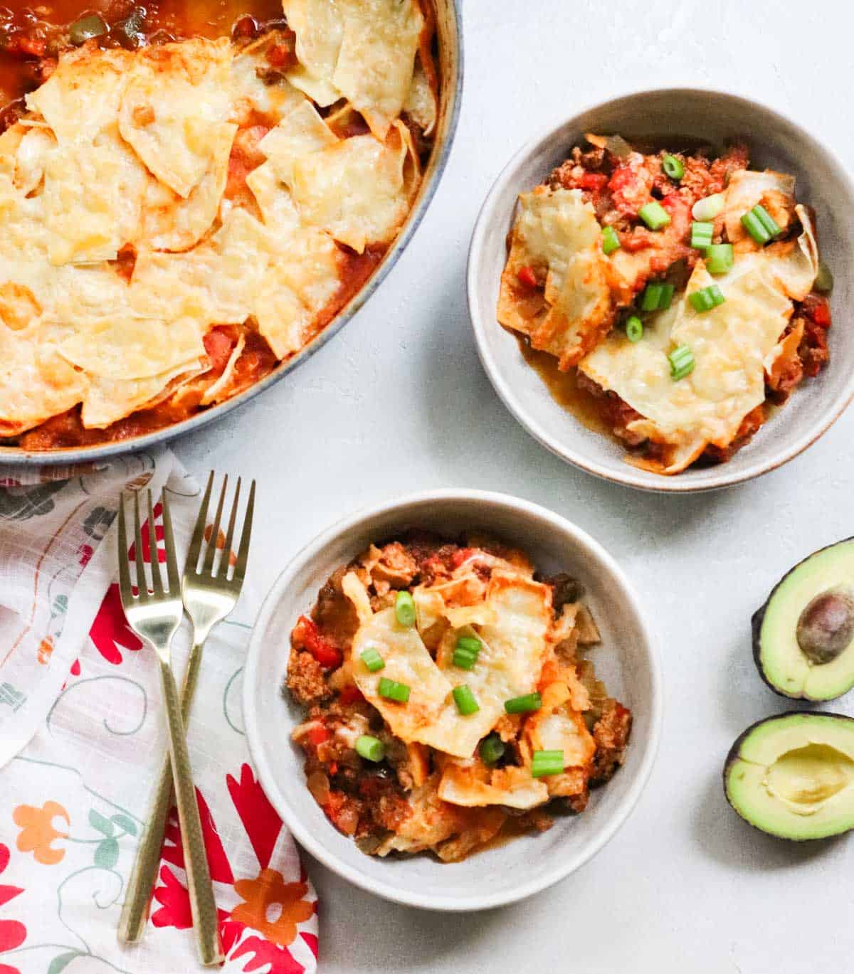 two bowls of taco casserole topped with chopped green onion, two gold forks, patterned napkin, fresh avocado, and pot with taco casserole.
