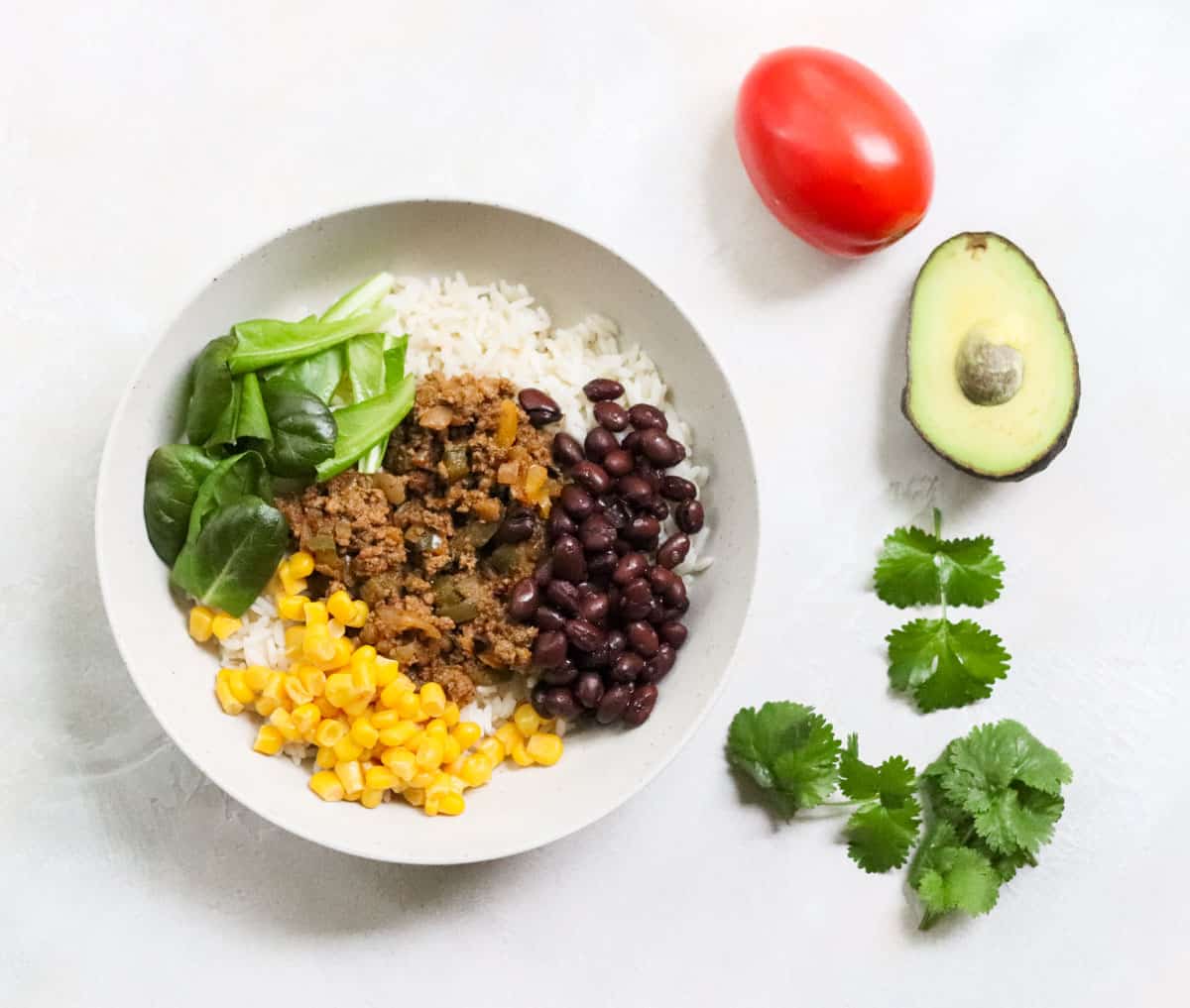 bowl with rice, black beans, taco meat, yellow corn, and lettuce next to a cut avocado, tomato, and fresh cilantro.