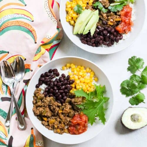 two ground beef taco bowls with various toppings next to a patterned napkin, cut avocado, silver spoons, and fresh cilantro.