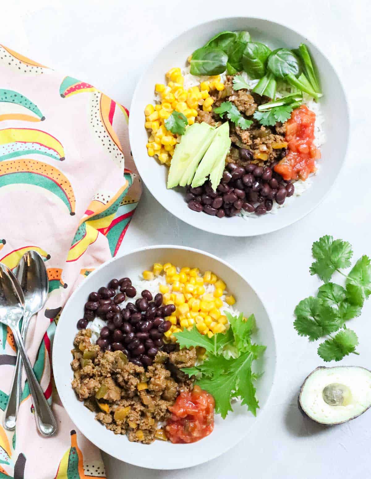two bowls filled with rice, taco meat, salsa, lettuce, corn, and beans next to a banana patterned dish towel, avocado and cilantro with two silver spoons.