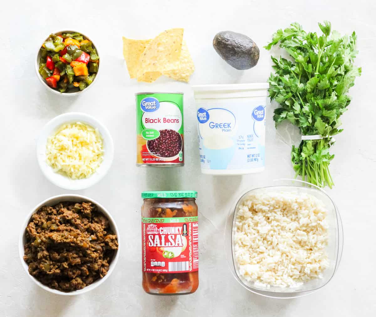 white counter with ingredients to make taco bowls including leftover taco meat, grated cheese, bell peppers, tortilla chips, black beans, salsa, rice, greek yogurt, cilantro, and an avocado.