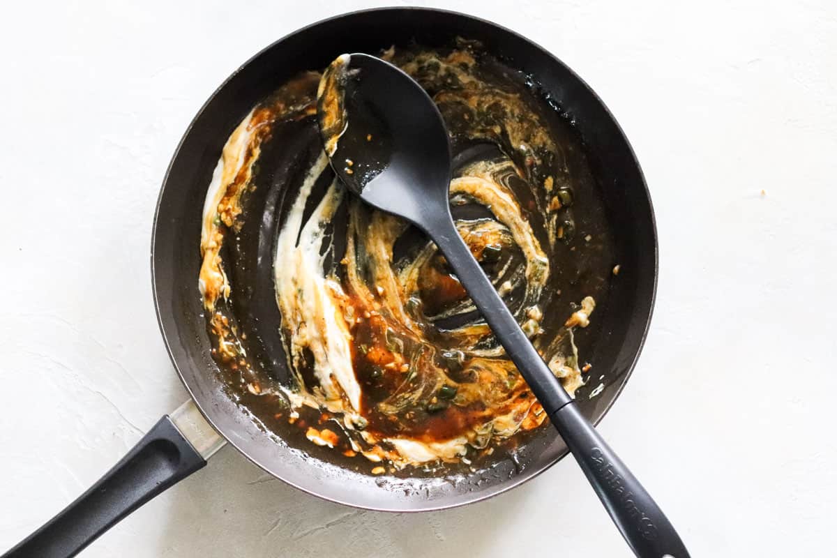 yogurt being stirred into gravy in a black skillet with a large spoon.