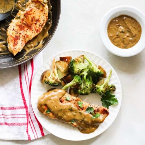 white plate with satuéed chicken and gravy with roasted potatoes and broccoli next to a white and red napkin, skillet with chicken, and bowl of extra homemade gravy.