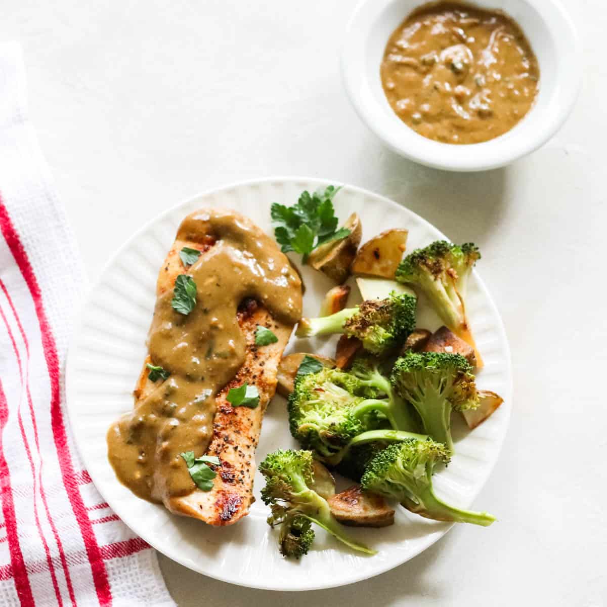 plate of chicken, homemade gravy, and roasted potatoes and broccoli next to a bowl of extra gravy and a red and white napkin.