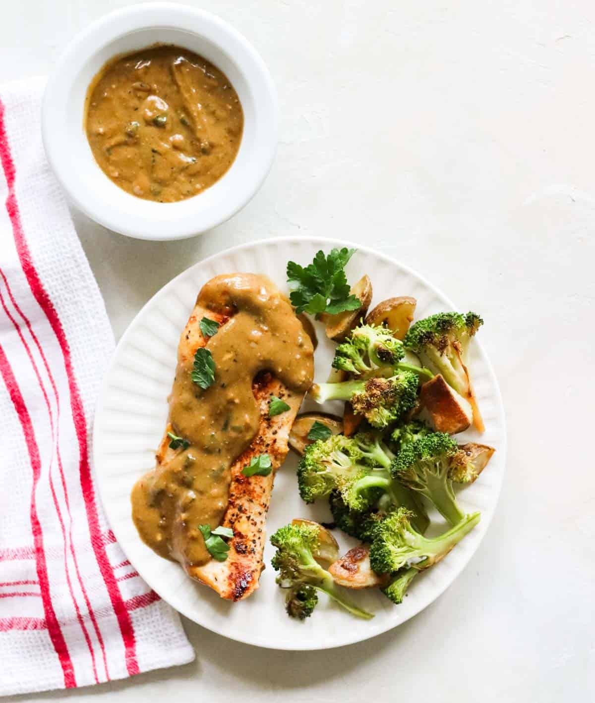 red and white napkin under a plate of chicken, homemade gravy, and roasted potatoes and broccoli next to a bowl of extra gravy.