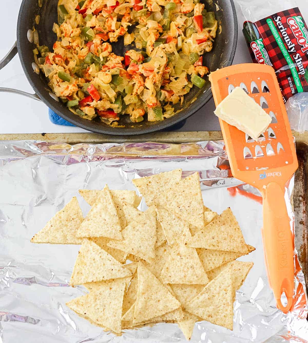 skillet with crawfish and veggies next to a baking sheet lined with foil covered in tortilla chips with an orange cheese grater and a black of cheddar cheese.